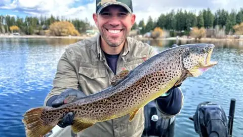Man proudly holding a freshly caught brown trout on a boat in a scenic lake, with a background of trees and cloudy sky.