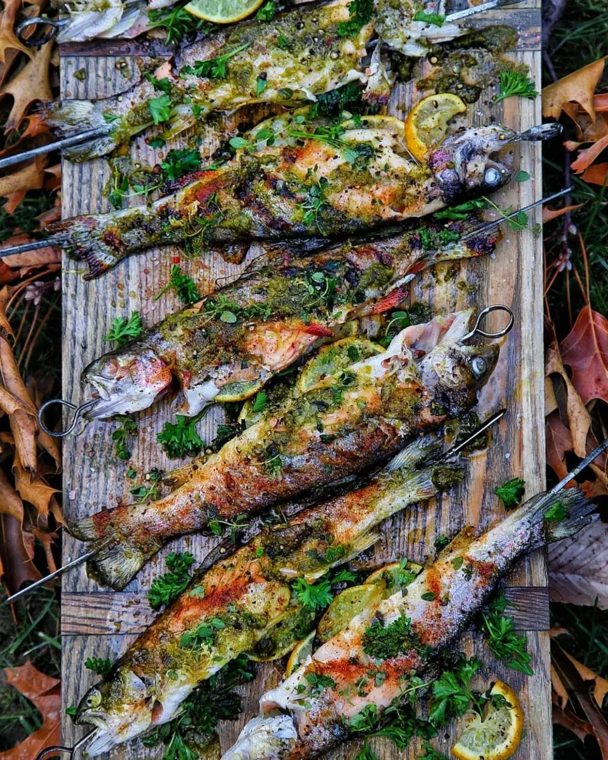 Grilled fish with herbs and lemon slices on a wooden platter, surrounded by autumn leaves.