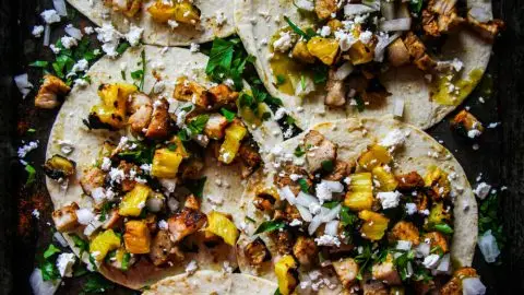 Delicious tacos al pastor with grilled pineapple, onions, cilantro, and crumbled cheese on a tray.