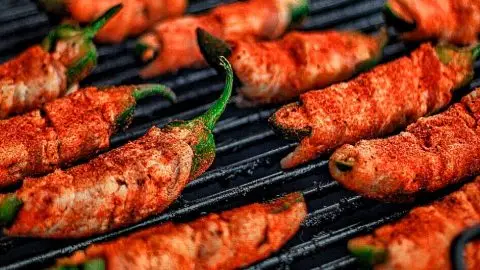 Grilled bacon-wrapped jalapeño poppers sizzling on a barbecue, seasoned with spices for a smoky flavor.