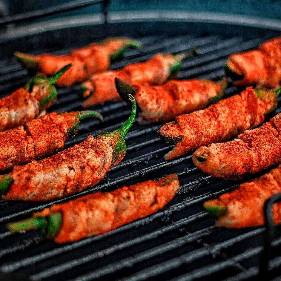 Grilled bacon-wrapped jalapeño poppers sizzling on a barbecue, seasoned with spices for a smoky flavor.