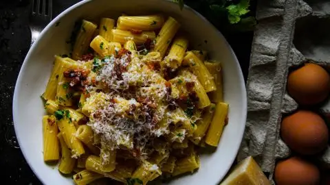 Creamy rigatoni carbonara with crispy bacon and cheese in a white bowl, surrounded by fresh parsley and eggs.