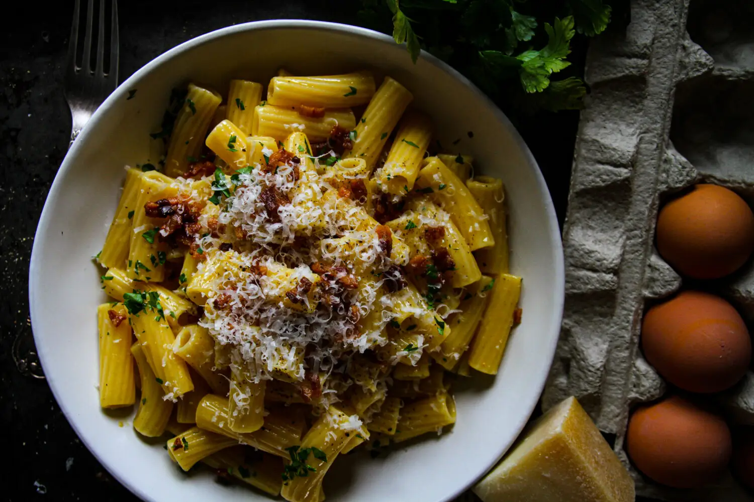 Creamy rigatoni carbonara with crispy bacon and cheese in a white bowl, surrounded by fresh parsley and eggs.