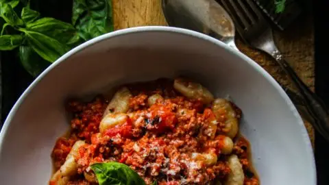 Plate of gnocchi bolognese garnished with basil, surrounded by fresh tomatoes and herbs.