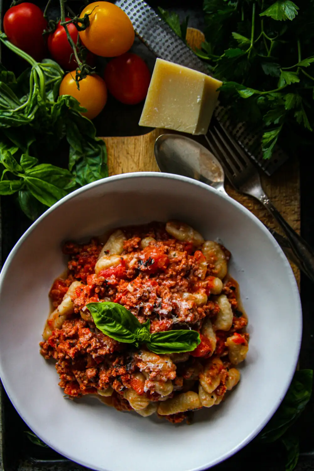 Plate of gnocchi bolognese garnished with basil, surrounded by fresh tomatoes and herbs.