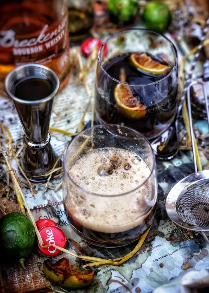 Cocktail ingredients: bourbon, lime, Coke cap, and strainer on a rustic map background.