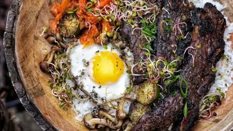 Delicious Korean bibimbap with grilled beef, fried egg, kimchi, mushrooms, and sprouts served in a rustic wooden bowl.