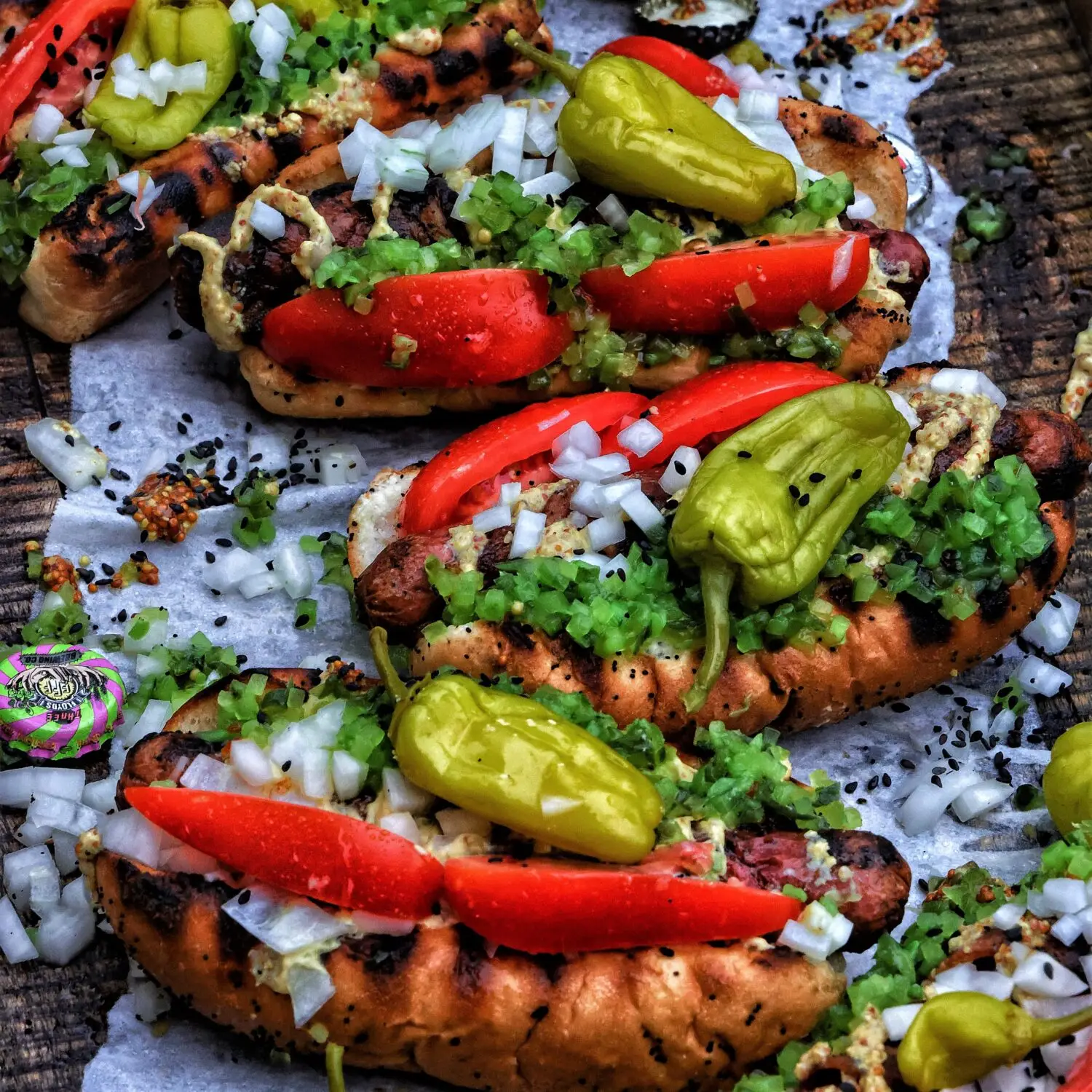 Grilled hot dogs topped with onions, pickles, tomatoes, and peppers on parchment paper. Delicious BBQ food.