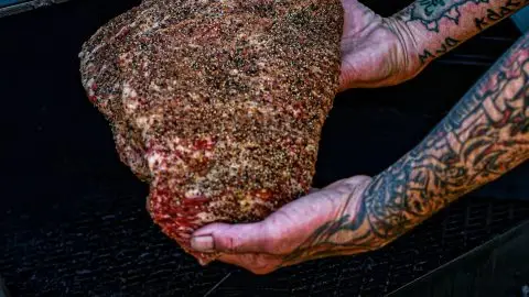Tattoed arms placing a seasoned brisket into a smoker for BBQ cooking.