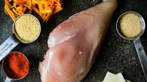 Raw chicken breast, broccoli, grated cheese, ham, Swiss cheese, and spices on a dark surface for cooking.
