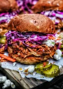 Pulled pork sandwich with vibrant coleslaw and pickles on a wooden board.