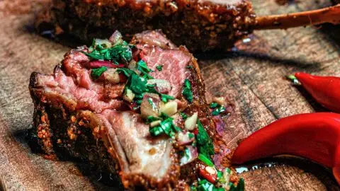 Grilled lamb chops garnished with fresh herbs on a rustic wooden board, accompanied by sliced red chili peppers.