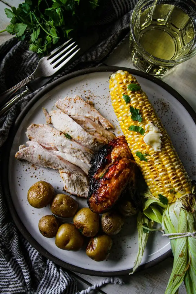 Grilled chicken with roasted potatoes, corn on the cob, and a glass of white wine on a plate with parsley garnish.