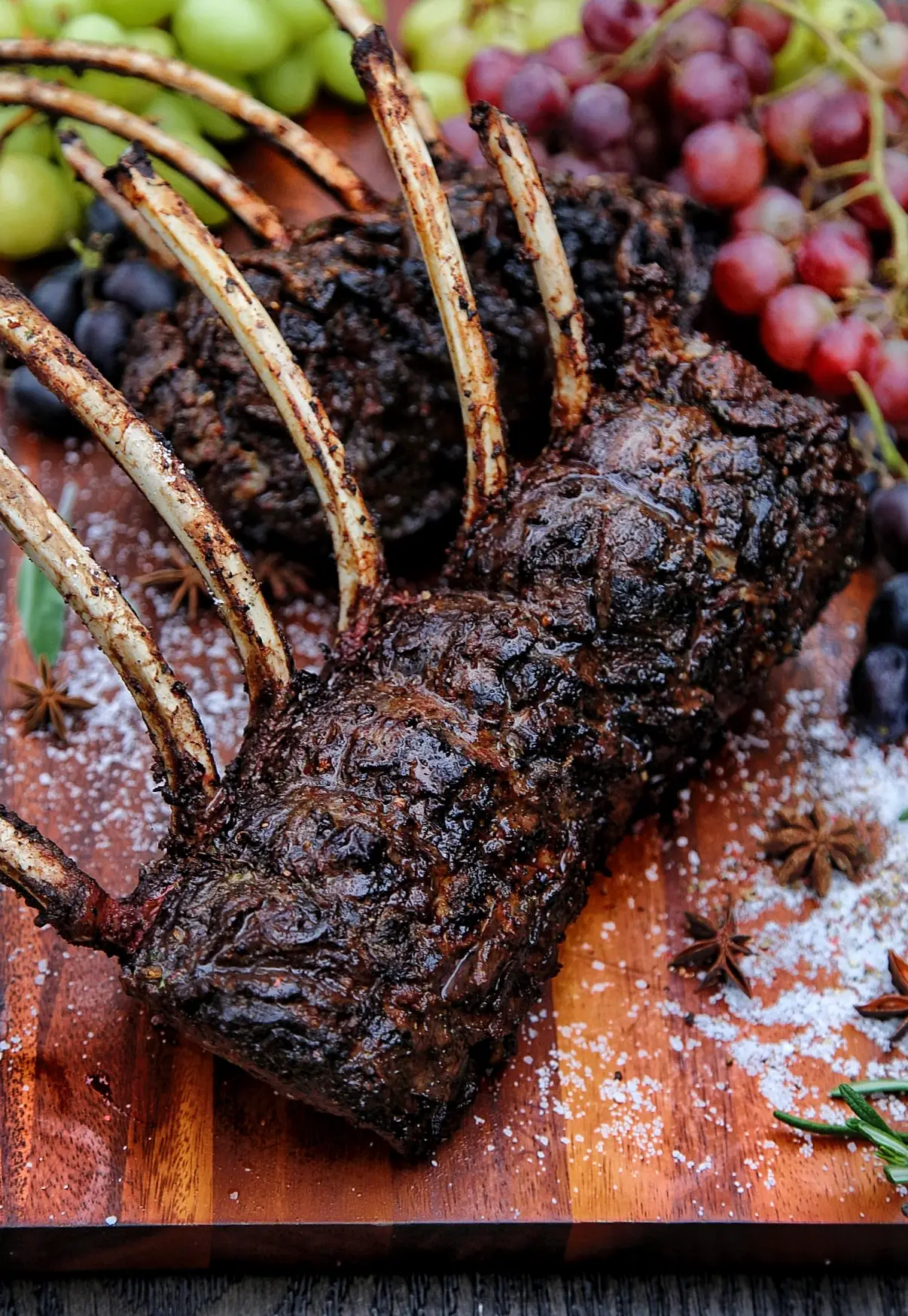 Grilled rack of lamb with juicy grapes on a wooden board, sprinkled with herbs and spices.