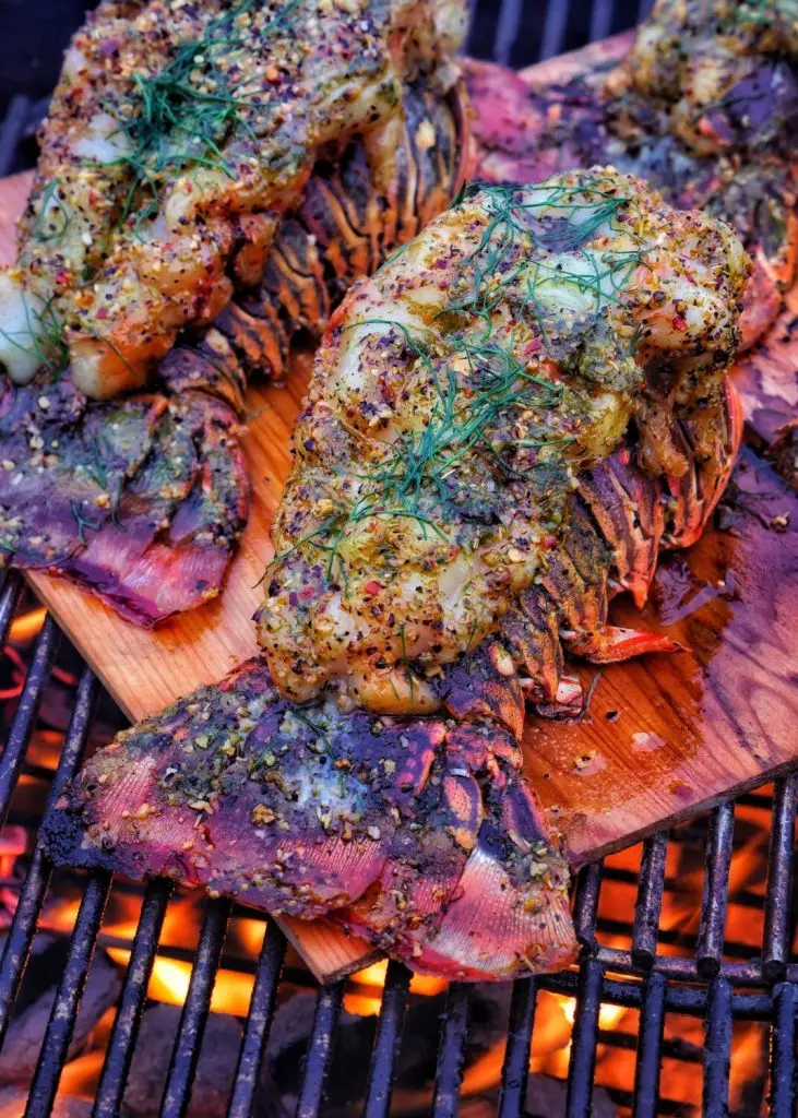 Grilled lobster tails on a cedar plank, seasoned with herbs and spices, with a fiery grill in the background.