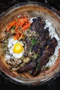 Delicious Korean BBQ beef bowl with fried egg, kimchi, mushrooms, sprouts, and rice on a rustic wooden plate.
