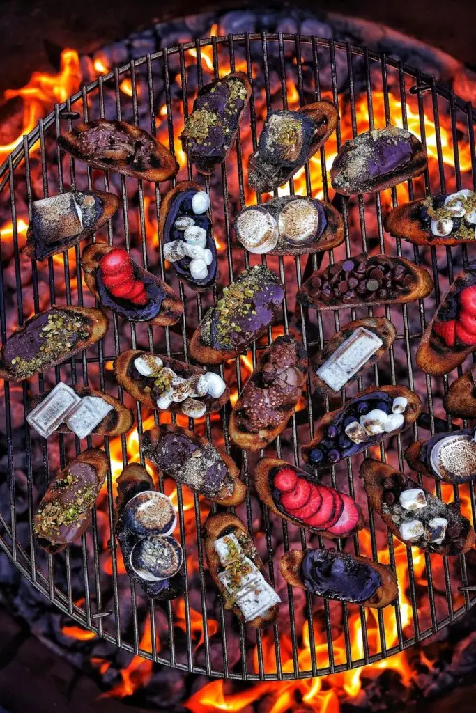 Grilled desserts with chocolate, berries, and marshmallows on toasts over flames, creating a delicious campfire treat.