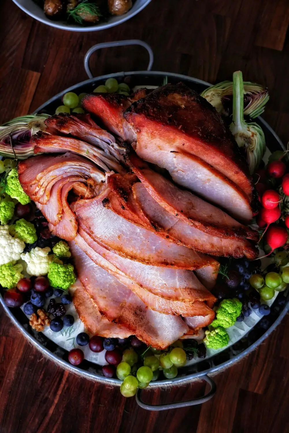 Sliced roast ham garnished with grapes, cauliflower, and blueberries on a serving platter.