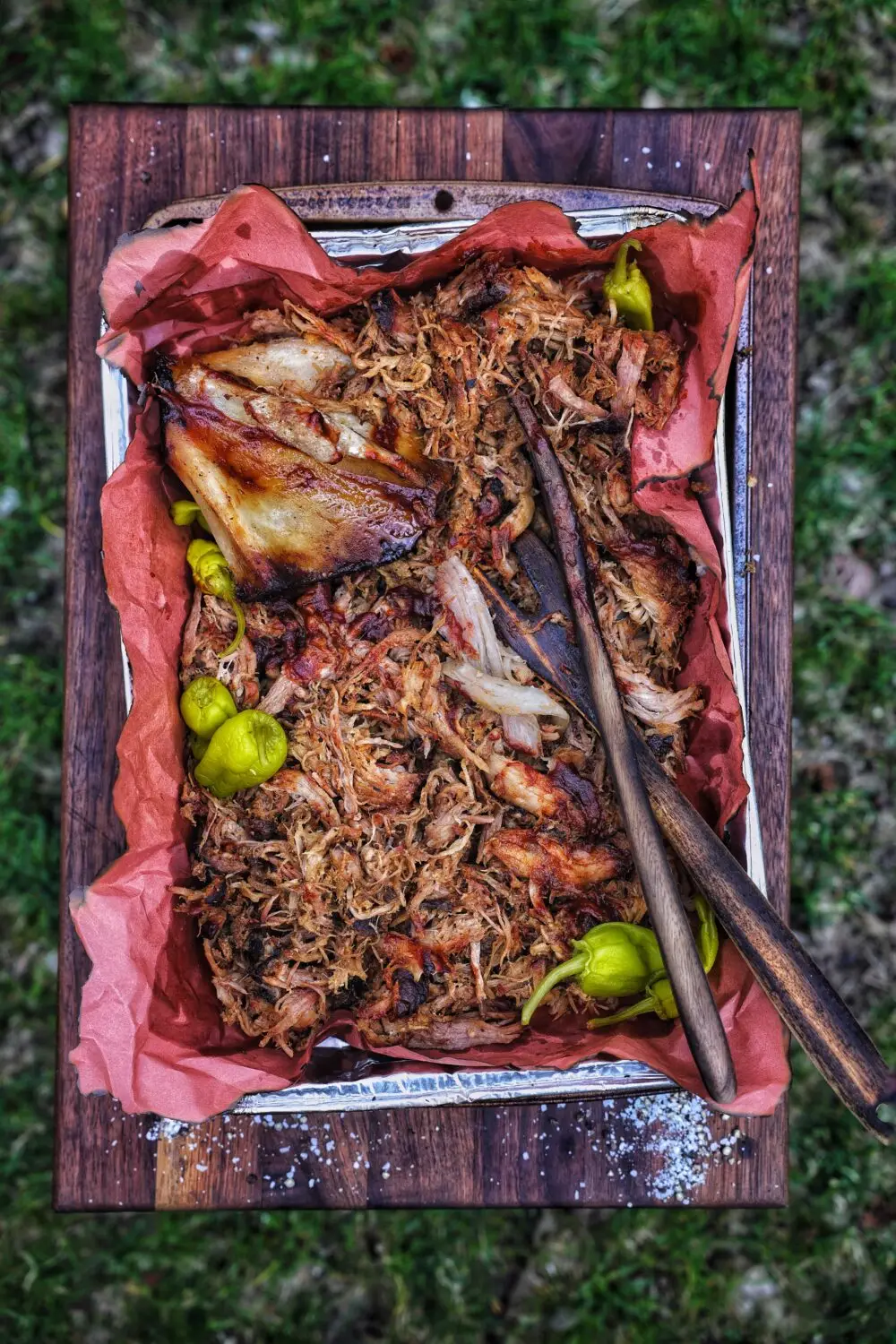 Slow-cooked pulled pork with roasted peppers on a rustic wooden tray, garnished with sea salt, served outdoors.