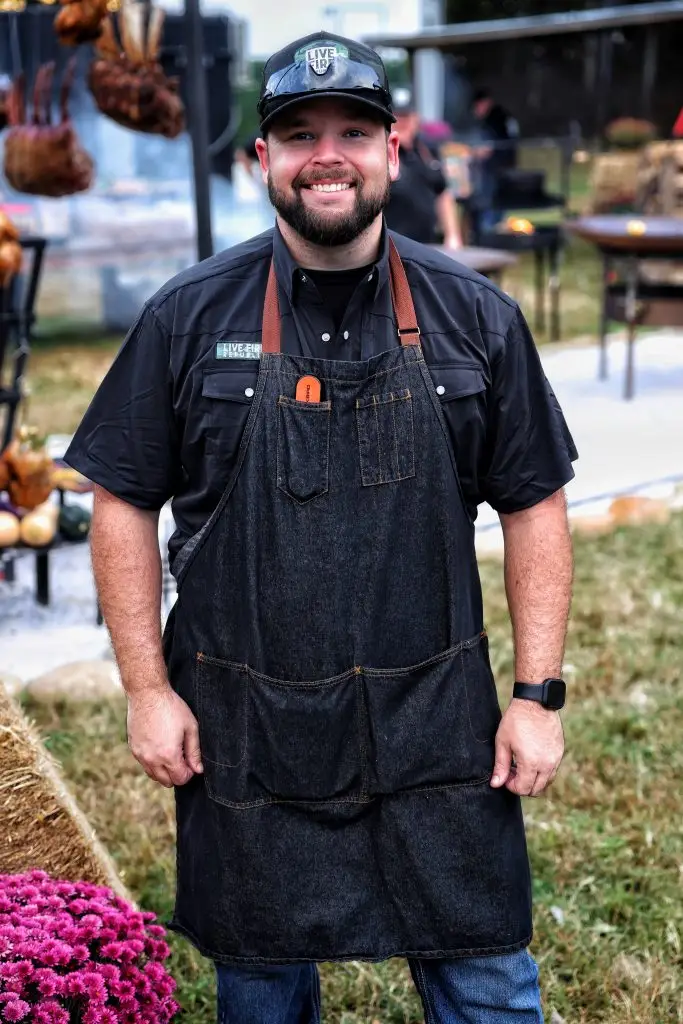 Man in apron and cap smiling at outdoor barbecue event. Grilled meats and smoke in the background.