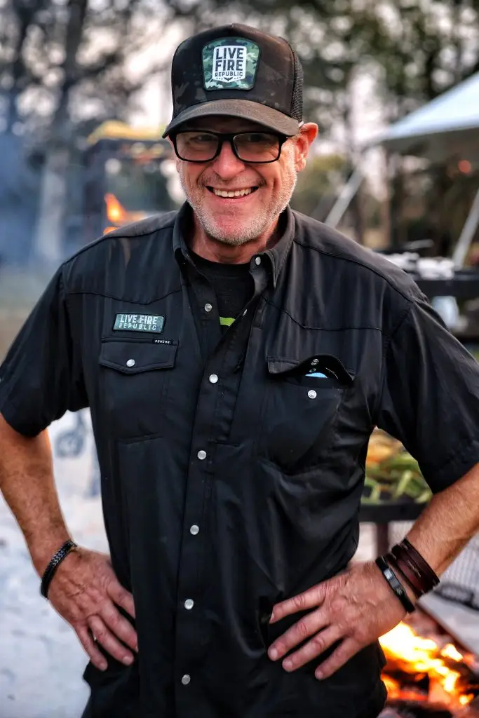 Man in black Live Fire Republic shirt and cap smiling outdoors near a fire.