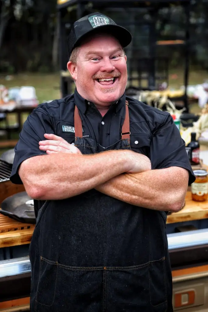 Smiling chef wearing a Live Fire Republic cap and apron, standing with crossed arms at an outdoor cooking event.