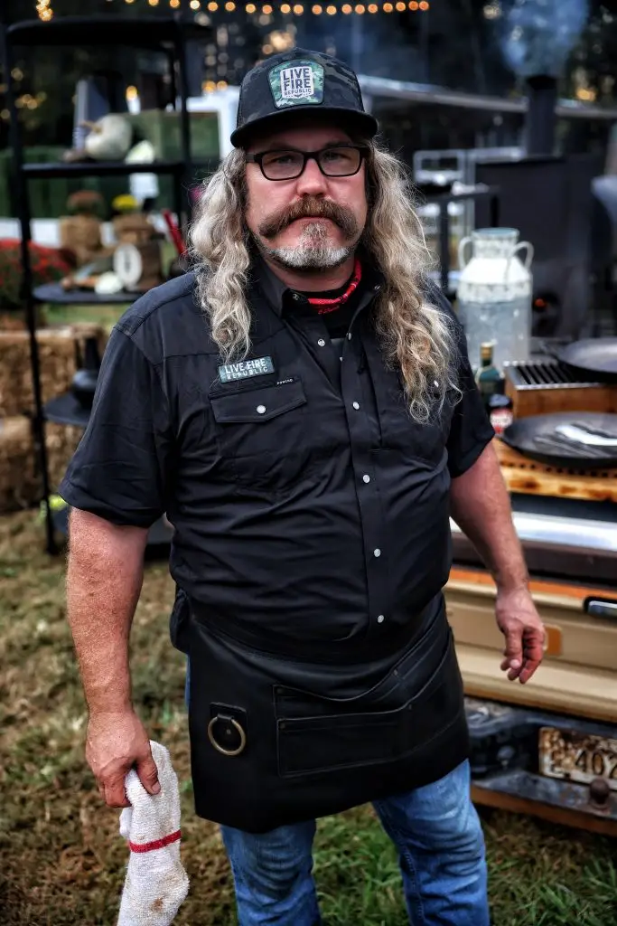 Man with long hair and beard in black chef attire at outdoor barbecue event, holding towel.