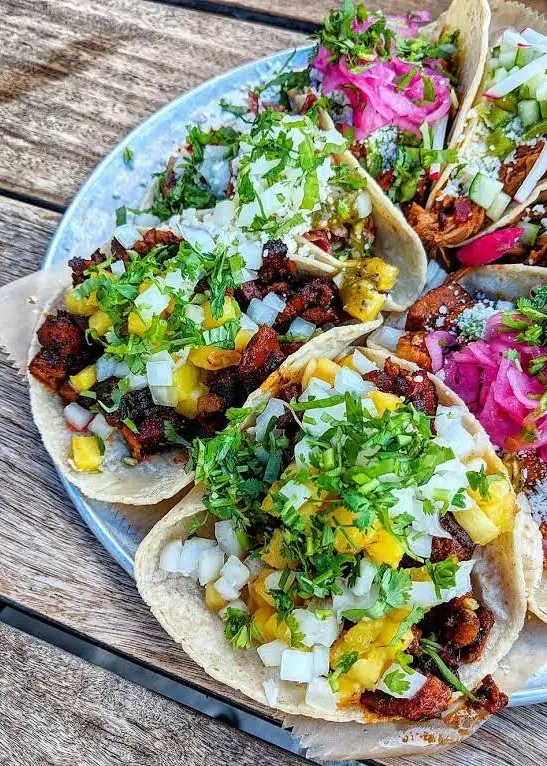 Plate of vibrant tacos with cilantro, onion, and pickled toppings on a wooden table, showcasing delicious street food.