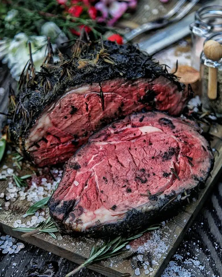 Juicy rosemary and pepper-crusted prime rib roast on wooden board with coarse salt and herbs.