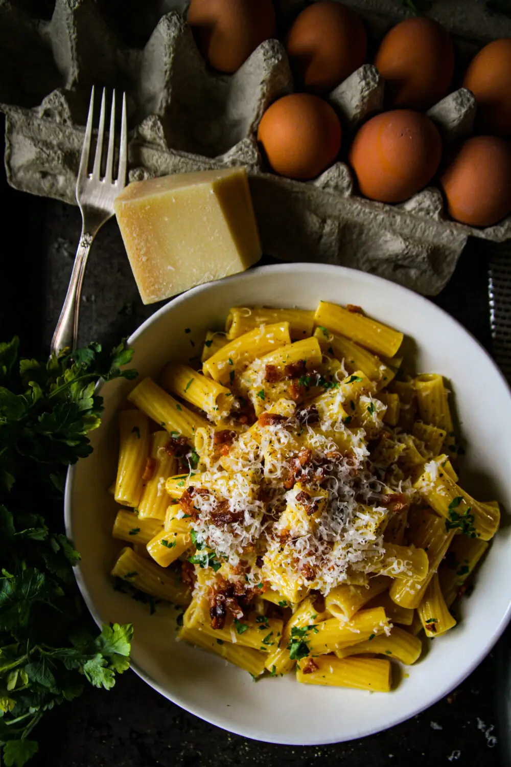 Delicious rigatoni carbonara topped with cheese, eggs, and bacon, surrounded by fresh parsley and whole eggs.