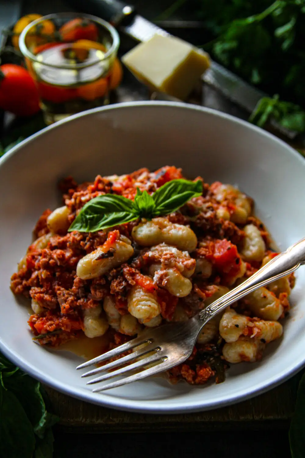 Plate of gnocchi with meat sauce and basil garnish, accompanied by a fork. Fresh ingredients in the background.