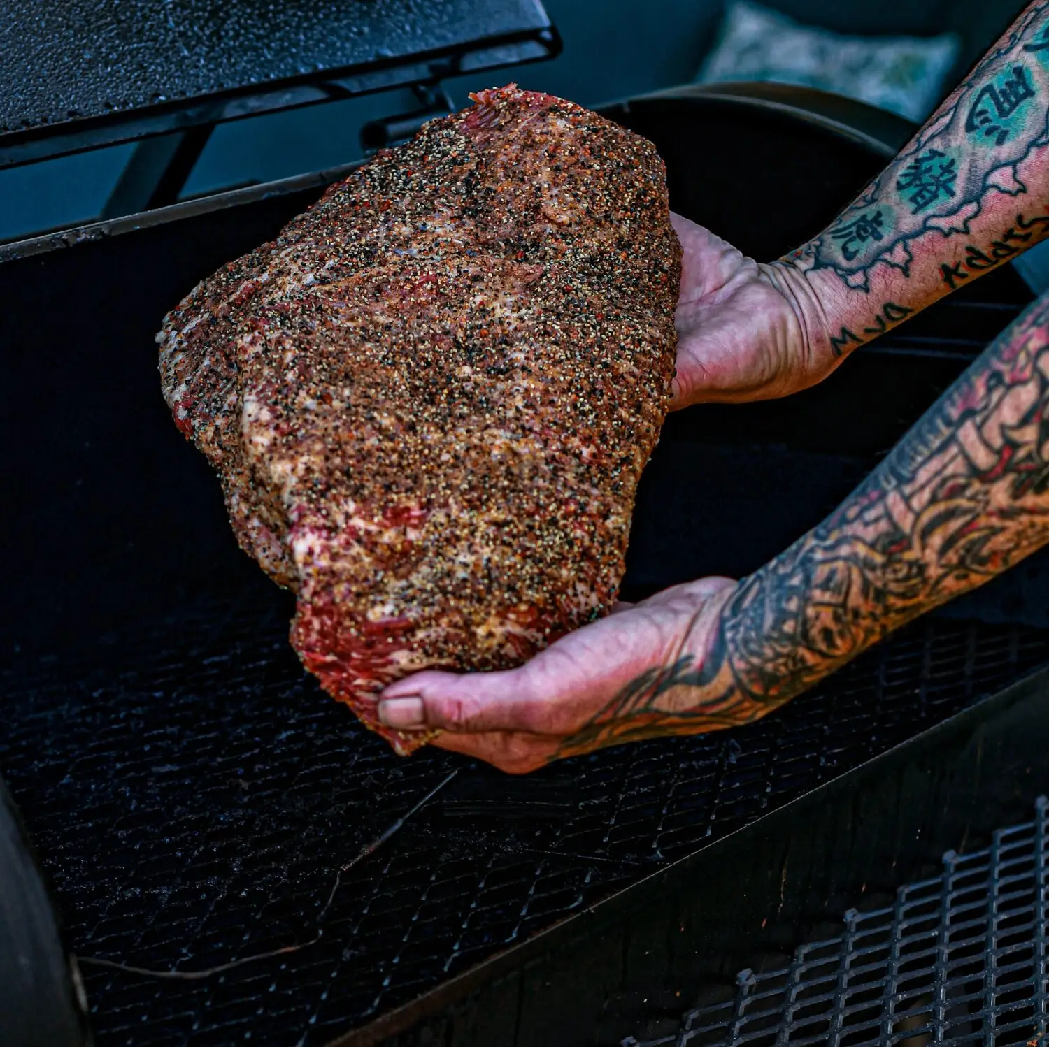 Tattooed hands hold a seasoned brisket above a smoker grill, ready for BBQ cooking.