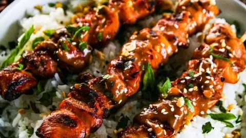 Grilled chicken skewers on rice with peanut sauce, garnished with sesame seeds and lime, next to cucumber salad.