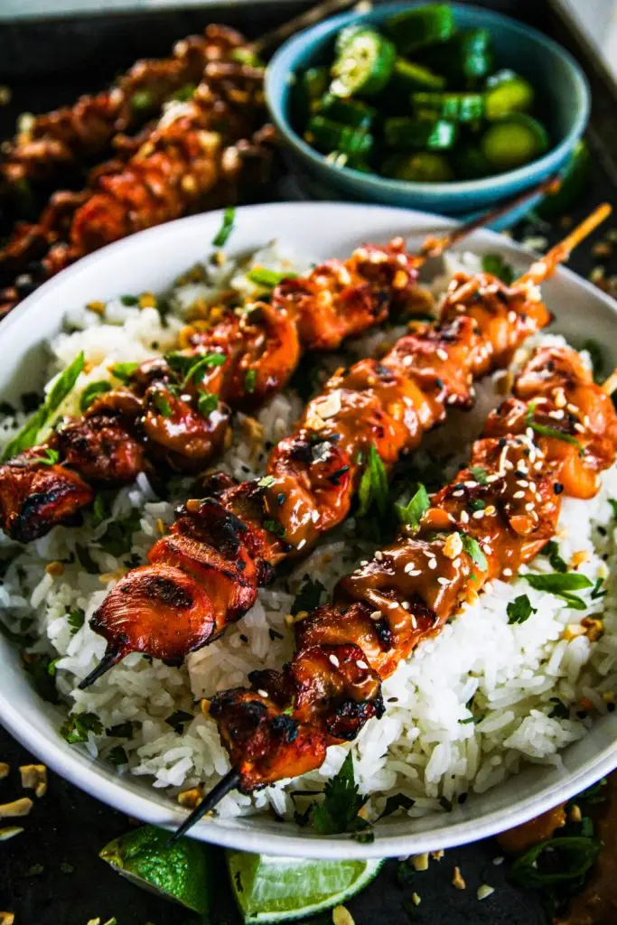 Grilled chicken skewers on rice with peanut sauce, garnished with sesame seeds and lime, next to cucumber salad.
