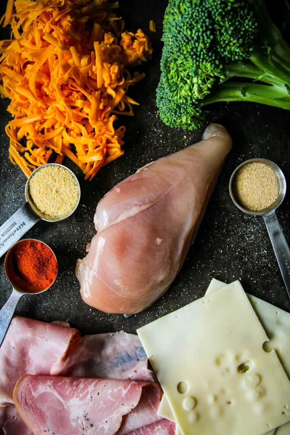 Ingredients for a flavorful chicken dish: raw chicken breast, broccoli, cheddar cheese, spices, and sliced ham.