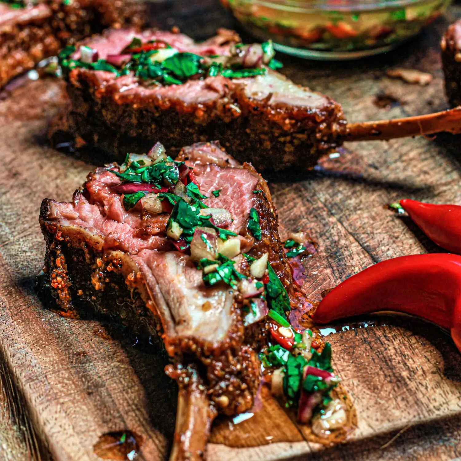 Grilled lamb chops garnished with herbs and spices, presented on a wooden board with red chili peppers.