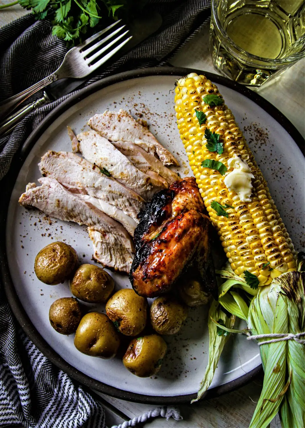 Grilled chicken and corn with roasted potatoes on a plate, garnished with herbs for a delicious summer meal.