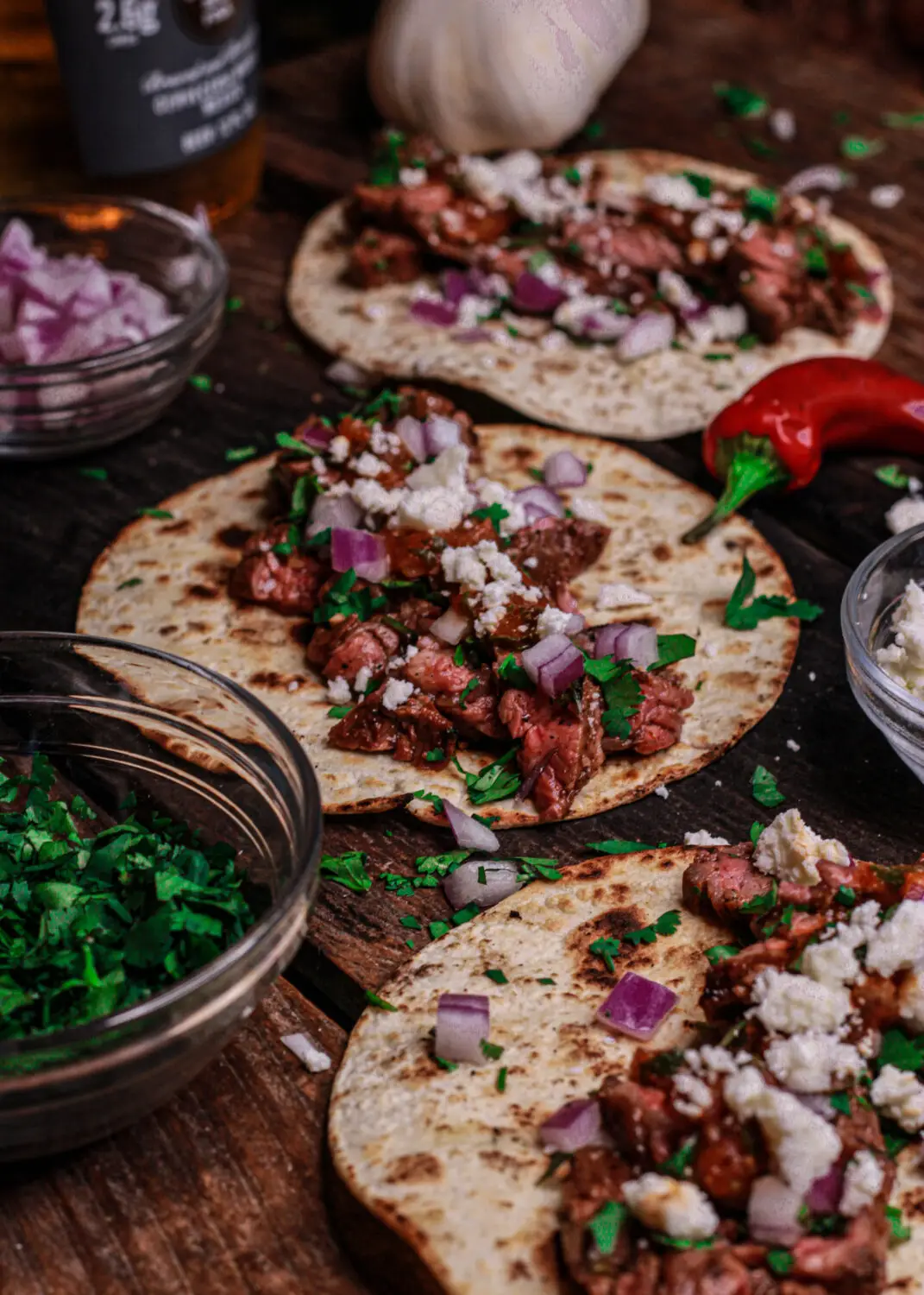 Street tacos topped with steak, onions, and cilantro on rustic wooden table with chili and ingredients.