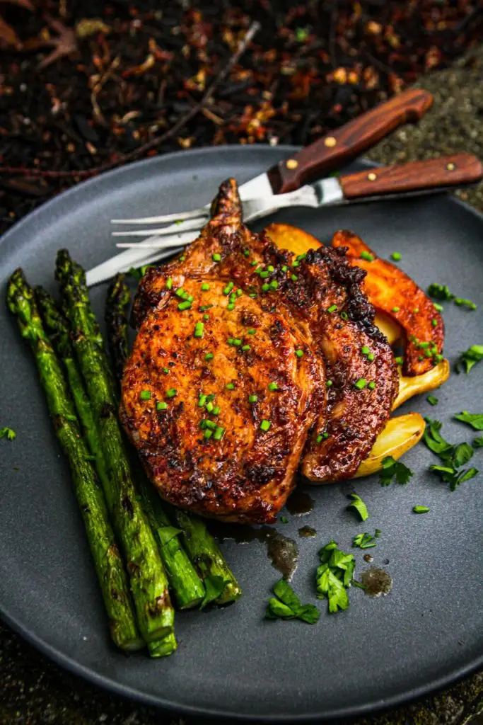 Grilled pork chop with chives, roasted potatoes, and asparagus on a gray plate for a delicious outdoor meal.
