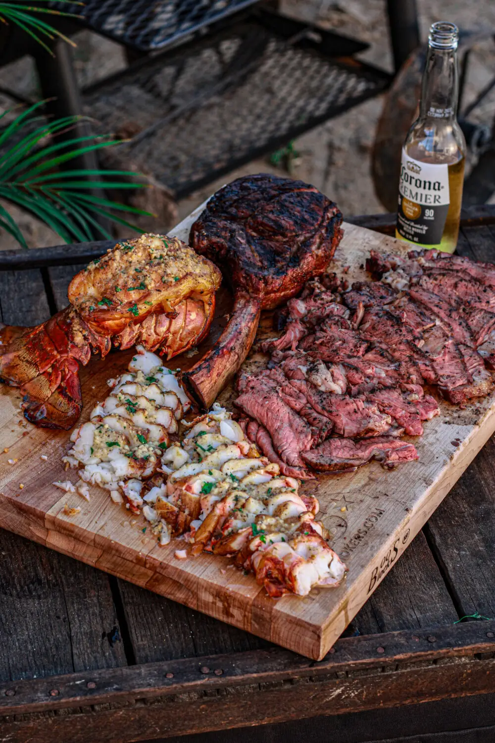 Grilled steak and lobster on a wooden board with a Corona Premier bottle in an outdoor setting.