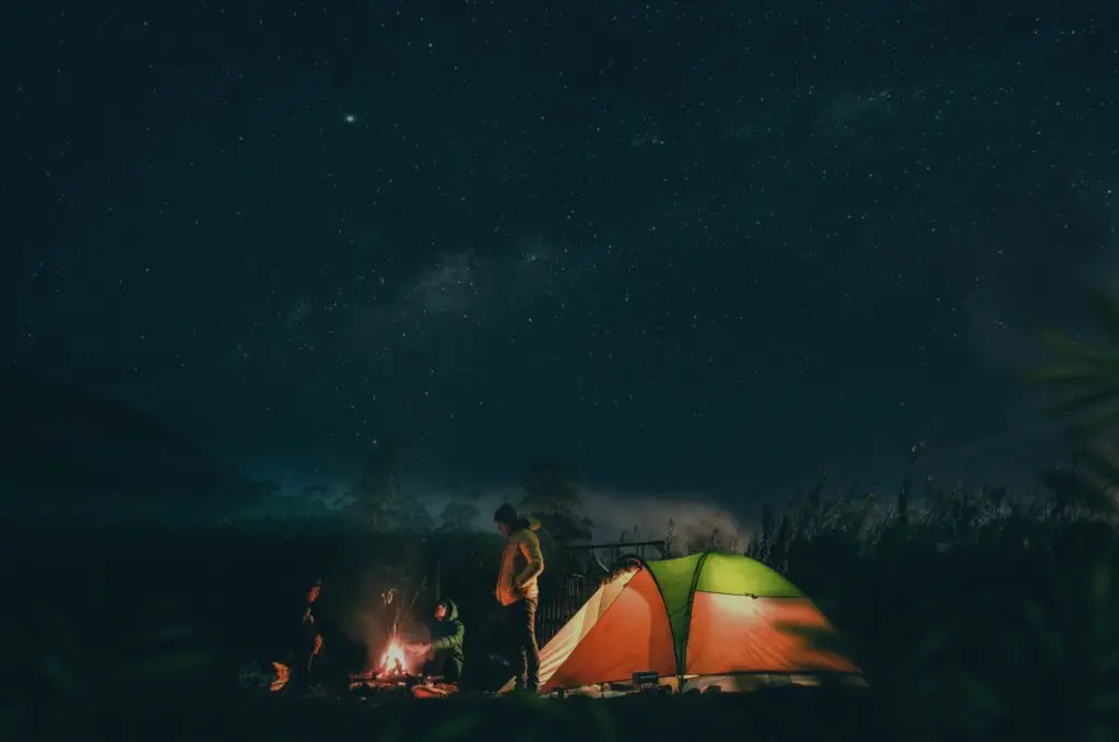 Camping under a starry night sky with a tent and campfire, surrounded by nature.