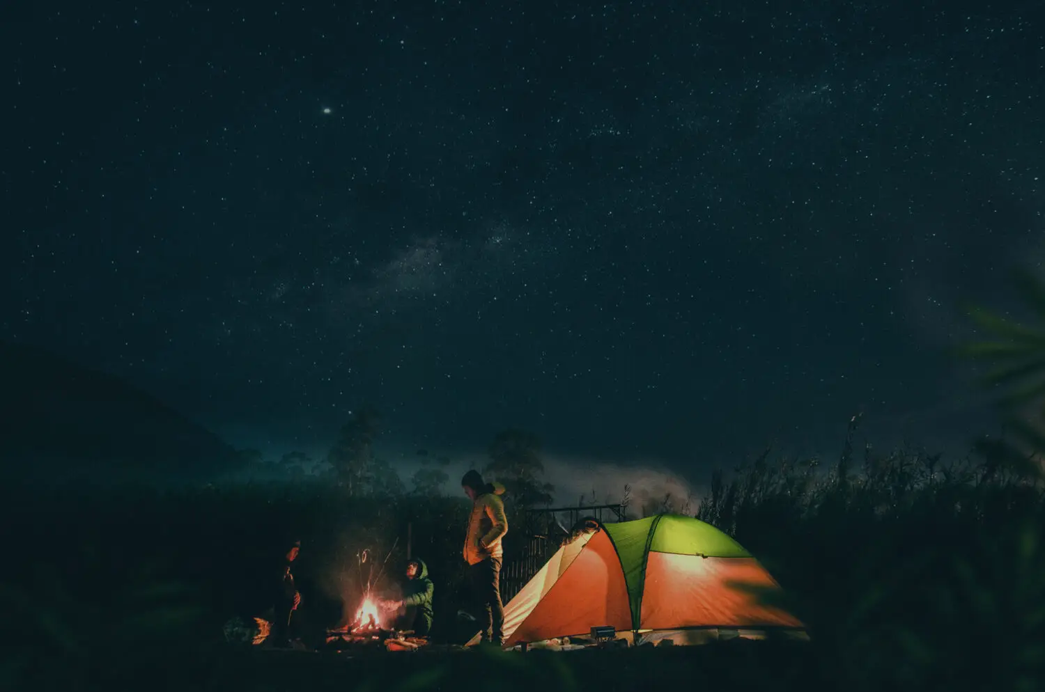 Camping under a starry night sky with a tent and campfire, surrounded by nature.
