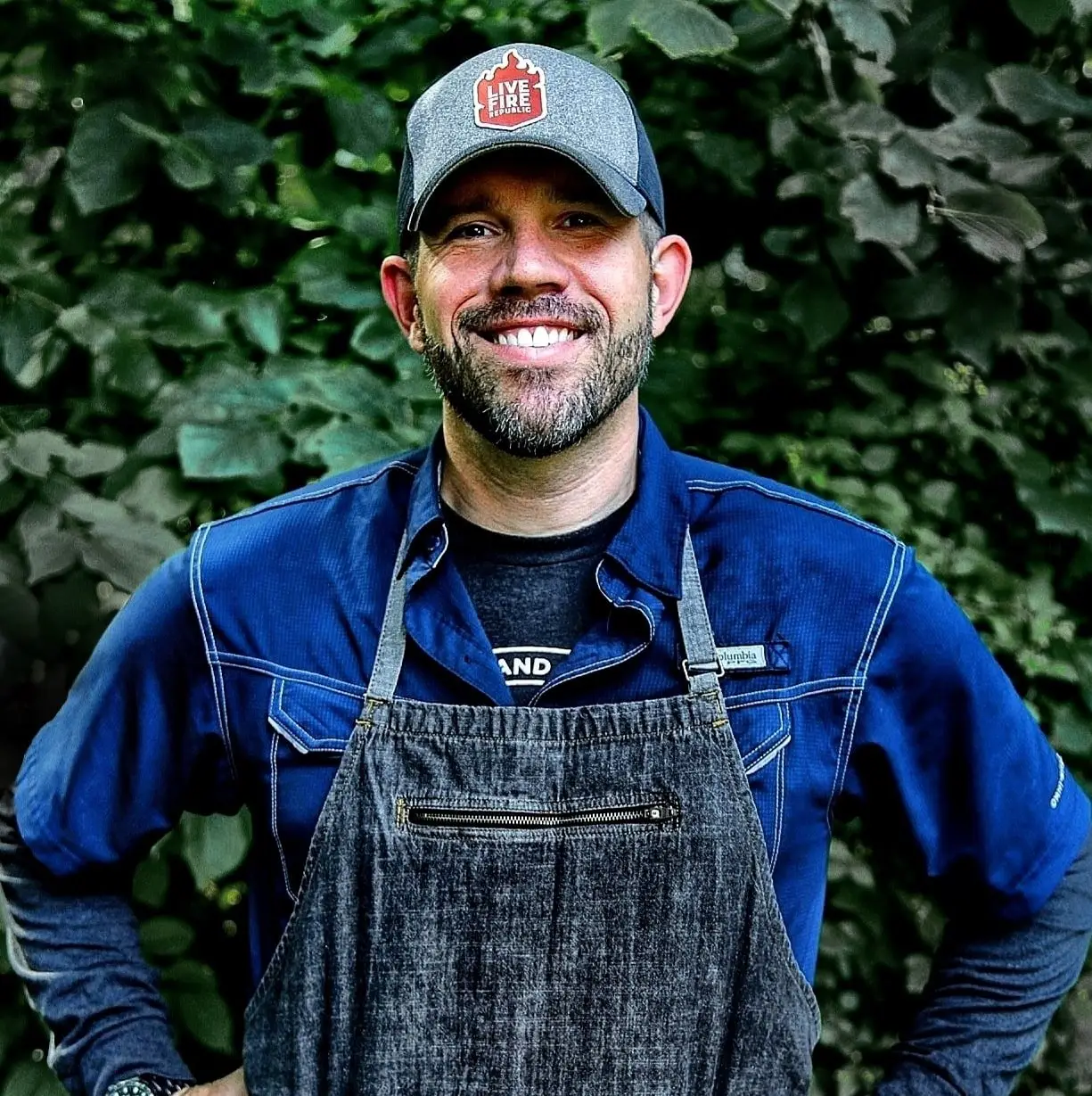 Smiling person wearing a cap and apron in front of green foliage. Casual outdoor setting.