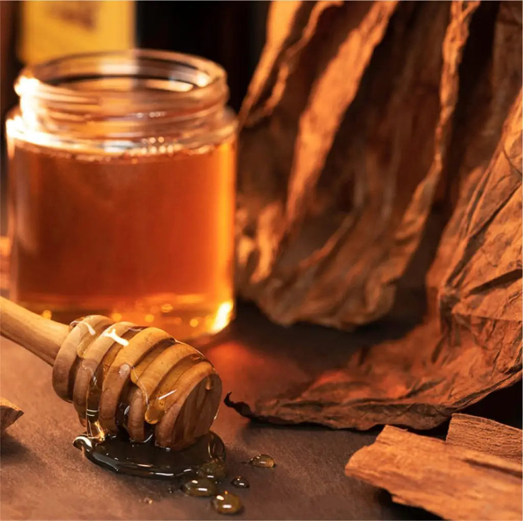 Honey jar with dipper surrounded by dried tobacco leaves on rustic table.