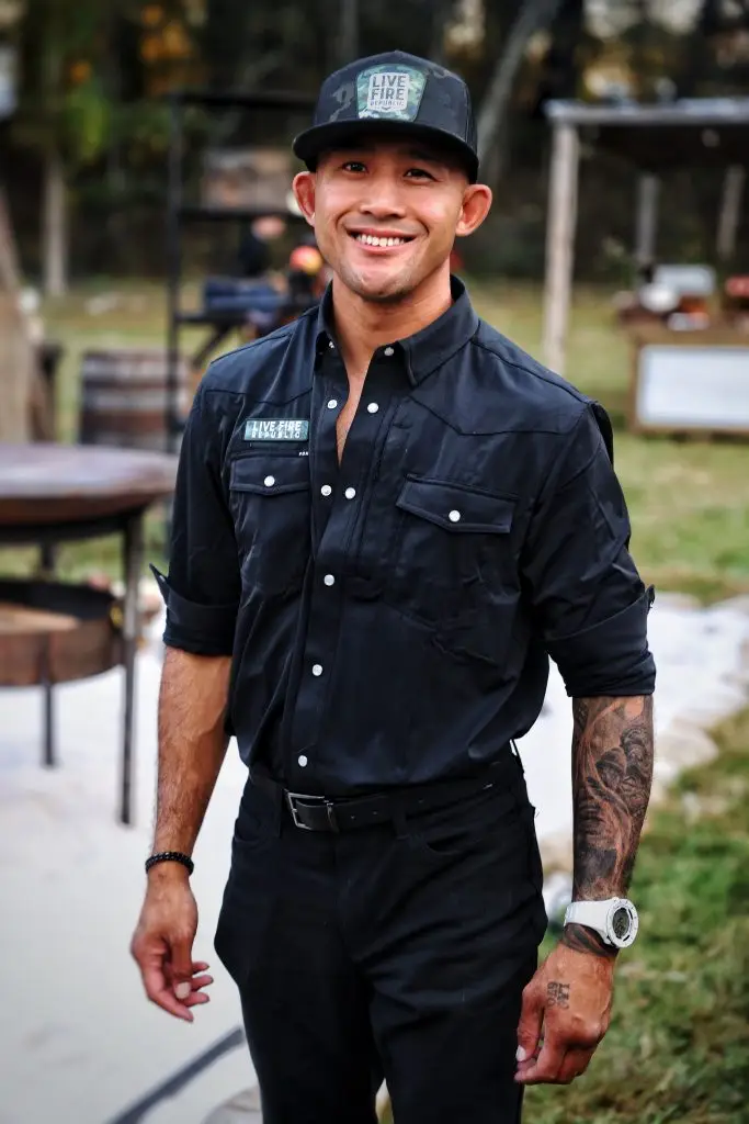 Smiling man in black shirt and hat with Live Fire Republic logo, standing outdoors with tattooed arm and watch.