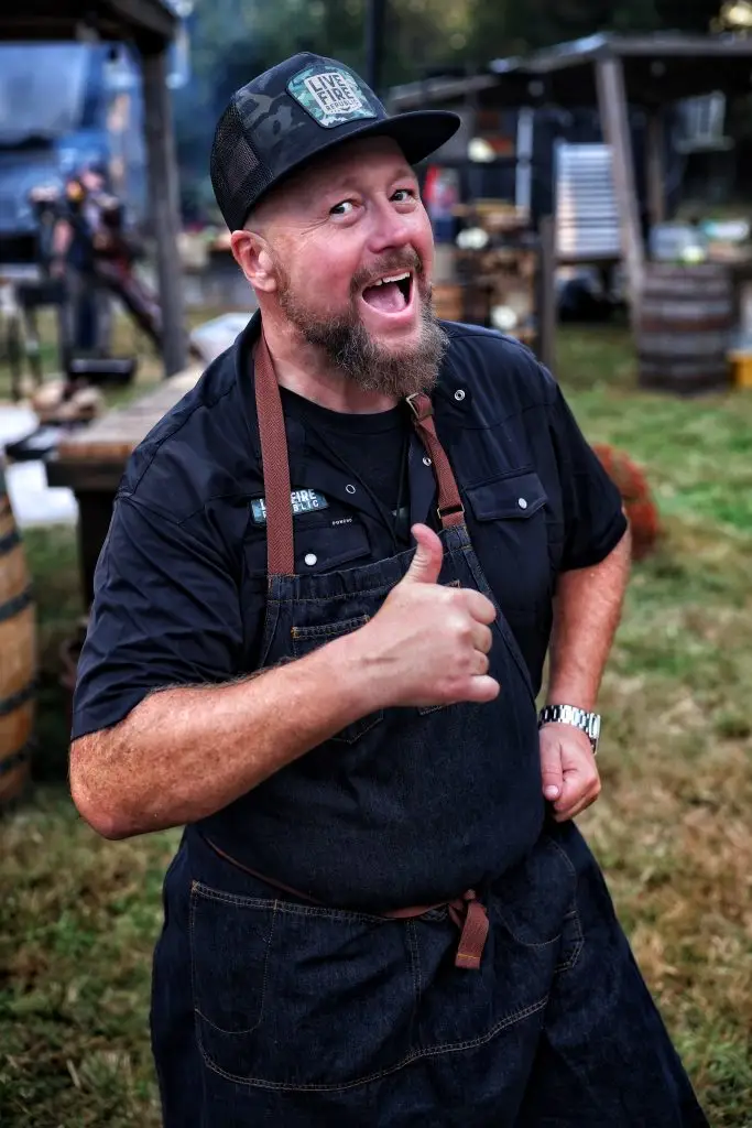 Cheerful man in an apron giving a thumbs-up at an outdoor event, wearing a hat with Live Fire logo.
