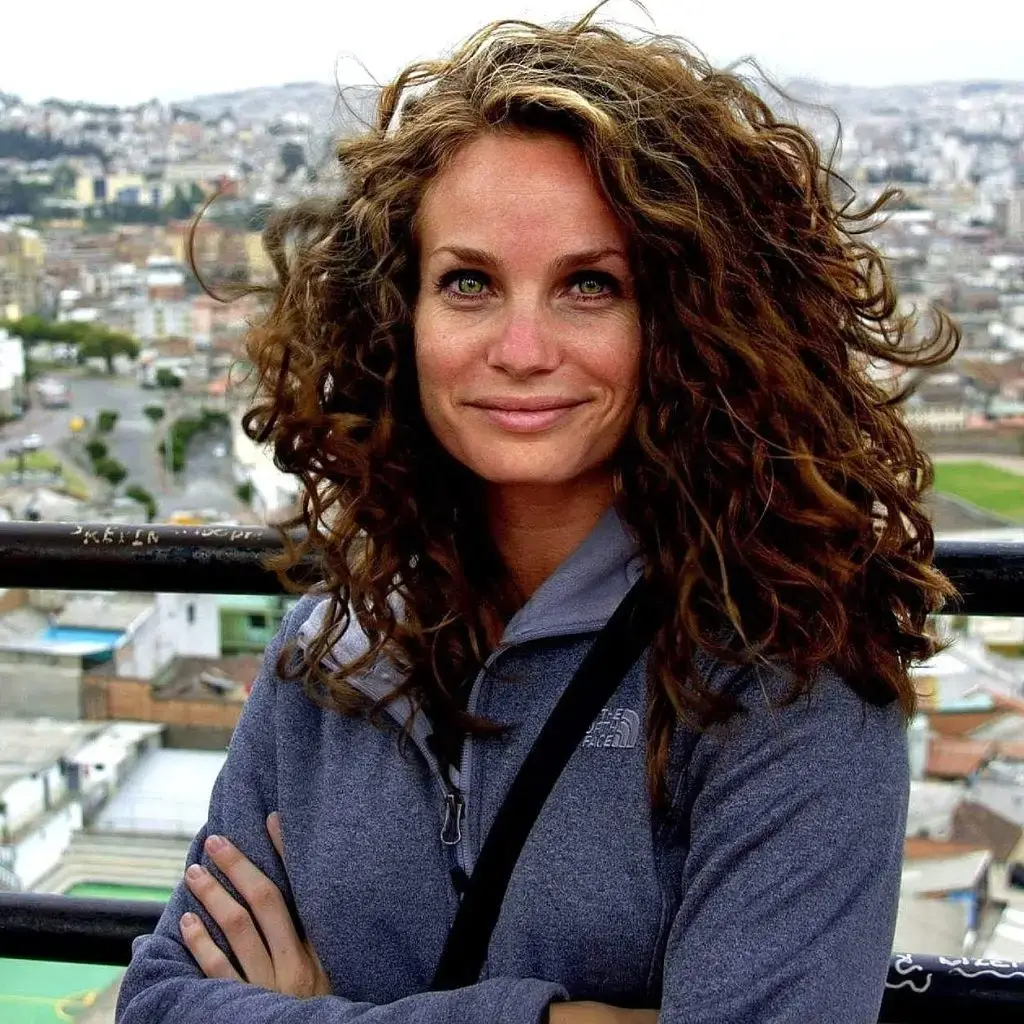 Woman with curly hair smiling on urban rooftop, wearing a gray jacket. Cityscape in the background, cloudy sky.