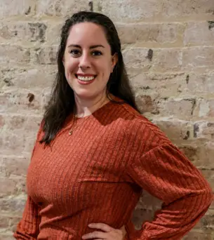 Smiling person in an orange sweater stands against a rustic brick wall background.