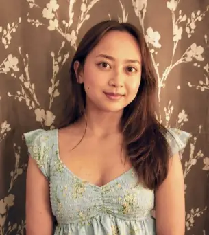 Woman in a floral dress standing against a patterned flower wallpaper backdrop, looking directly at the camera.